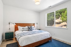 A bedroom with a wooden bed, white walls, a large window with a view of trees, and a green nightstand.