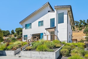 A modern two-story house with a concrete staircase, a wooden deck, and a landscaped garden.