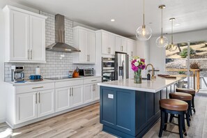 Una cocina moderna con gabinetes blancos, una isla azul, electrodomésticos de acero inoxidable y un amplio ventanal con vistas al exterior.