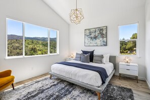 Un dormitorio con una cama grande, dos mesitas de noche, una silla y vistas a un bosque a través de amplios ventanales.