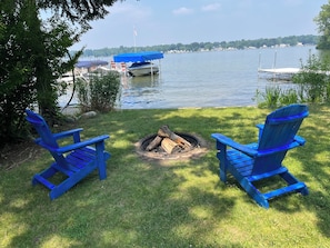 A lakeside view with two blue Adirondack chairs, a fire pit, and boats docked in the background. - Cozy Cottage Retreat on Jordan Lake with Amazing Views and Great Fishing (Lake Odessa)