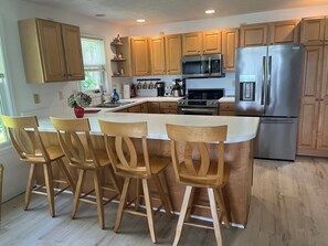 A kitchen with wooden cabinets, a stainless steel refrigerator, a microwave, and a sink. There are four wooden chairs around a central island. - Cozy Cottage Retreat on Jordan Lake with Amazing Views and Great Fishing (Lake Odessa)