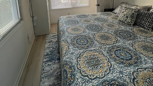 Ein Schlafzimmer mit Deckenventilator, einem Bett mit gemusterten Betttextilien, einem Fenster mit Jalousien und einer Tür.