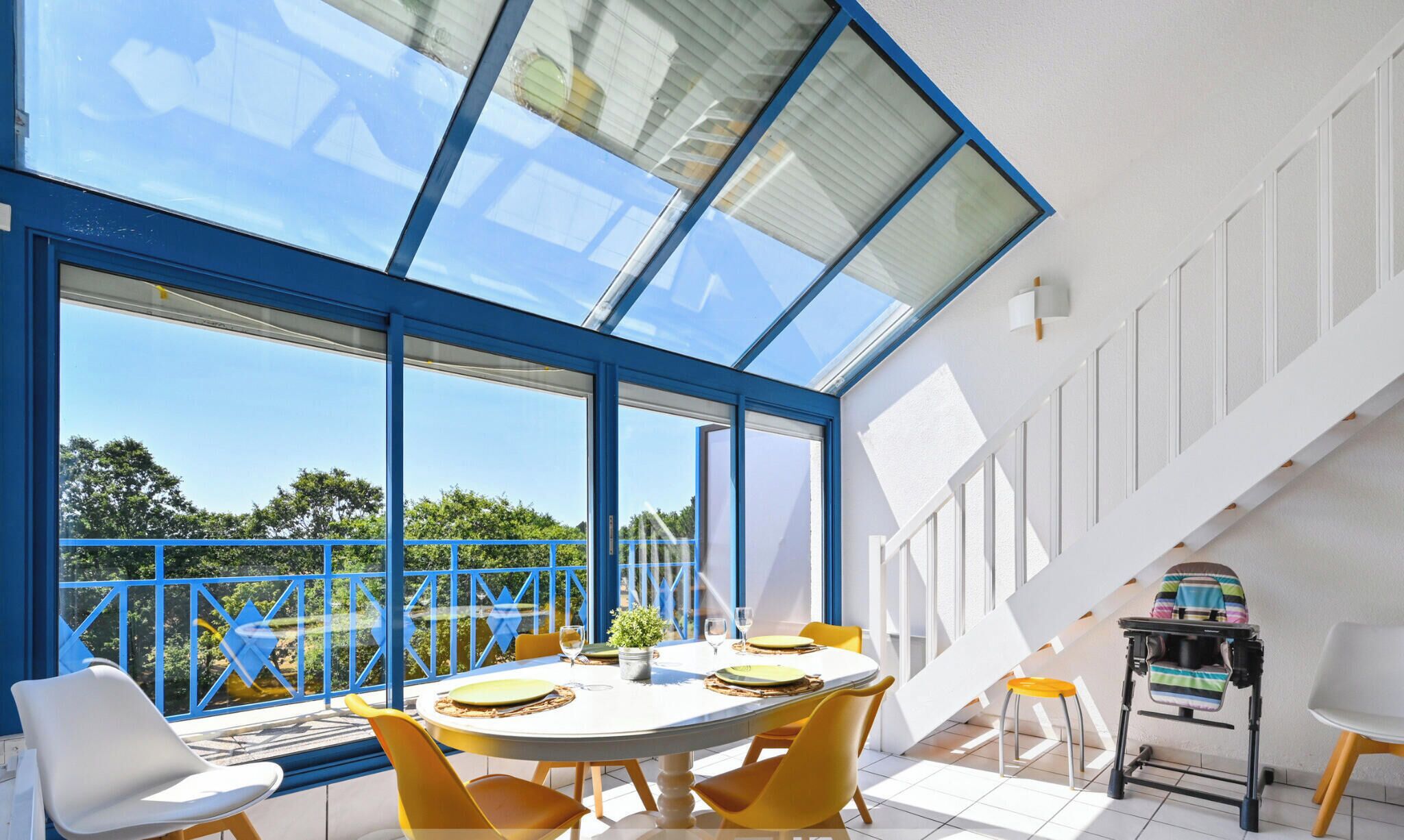 A modern dining area with a large table, yellow chairs, and a view through expansive windows.
