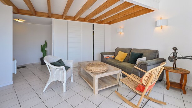 A living room with a wooden ceiling, a white wicker chair, a grey sofa with cushions, a small round table, and a decorative metal sculpture. - Le Scalby - Piscine - Golf (Pornic)