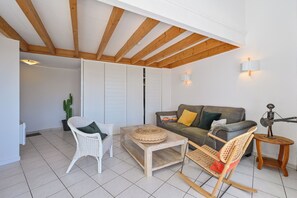 A living room with a wooden ceiling, a white wicker chair, a grey sofa with cushions, a small round table, and a decorative metal sculpture. - Le Scalby - Piscine - Golf (Pornic)