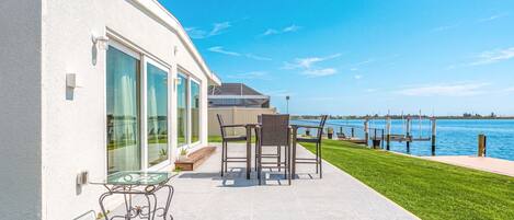 A modern house with a patio overlooking a body of water, featuring a glass table and chairs.