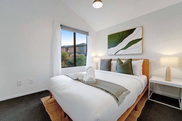 A hotel room with a large bed, a window with curtains, a bedside table with a lamp, and a wall-mounted artwork.