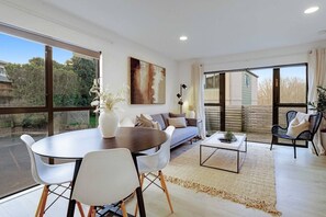 A modern living room with a dining area, large windows, and a view of the outdoors. - Vibrant Family 2BR Townhouse with Air-Con and Deck (Auckland South)