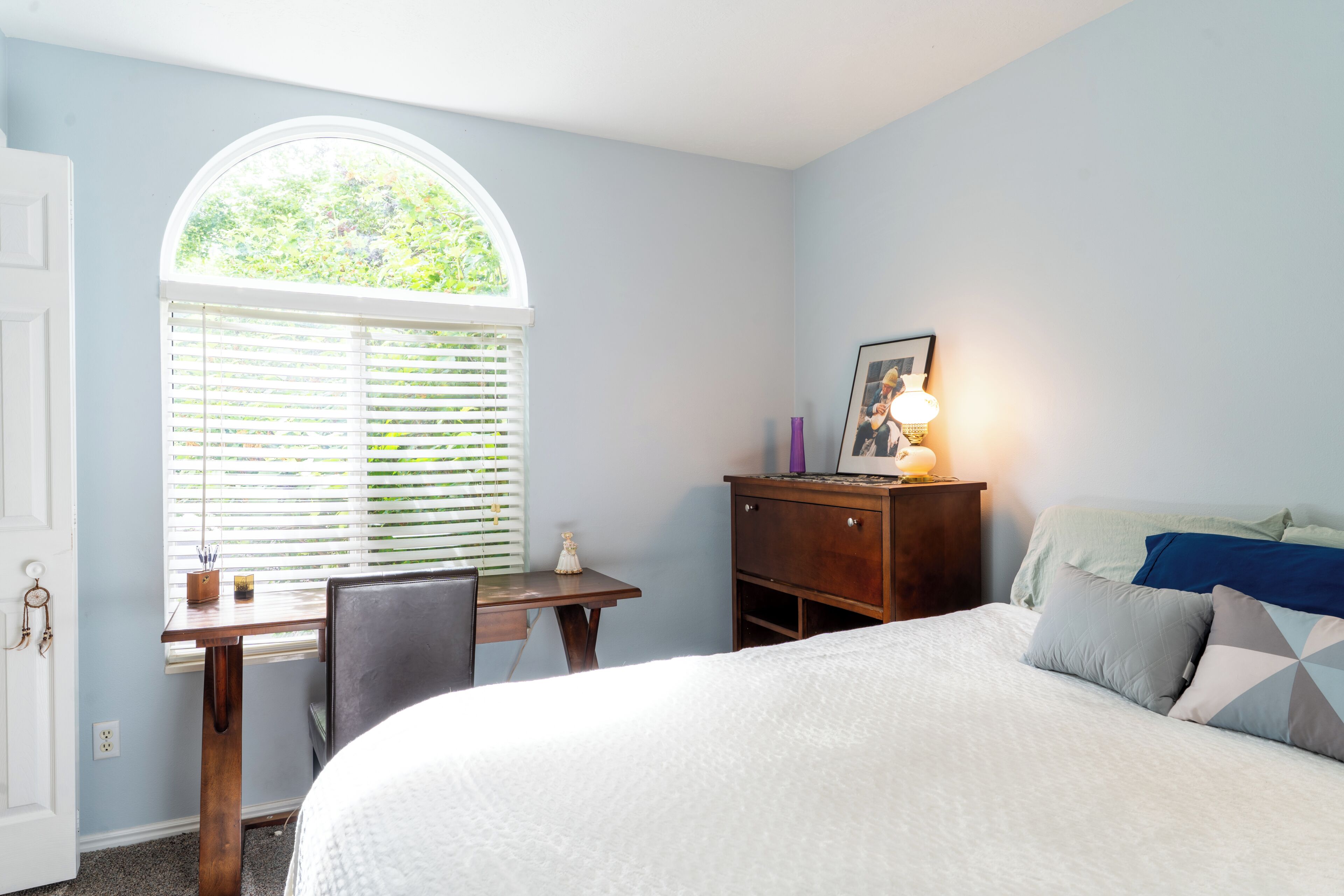 Une chambre à coucher comprenant un lit, un bureau, une chaise, une commode, une lampe et une fenêtre avec des stores.