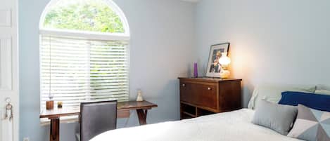 A bedroom with a bed, a desk, a chair, a dresser, a lamp, and a window with blinds.