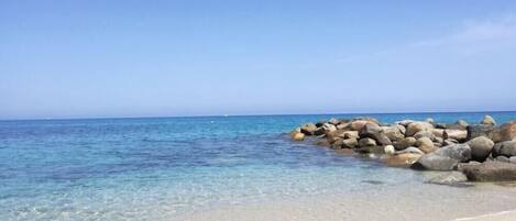A beach with clear water, a sandy shore, and a rocky outcrop.