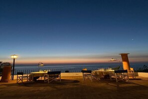Uma área de refeições ao ar livre com mesas, cadeiras, um candeeiro e vista para o mar ao entardecer.