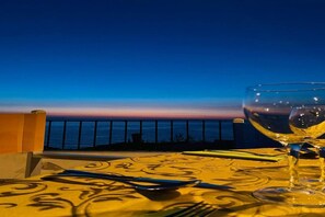A table set with a wine glass, cutlery, and a patterned tablecloth, overlooking a coastal view during twilight. - Residenza Villa Spagnola Adults Only (Parghelia)