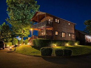 A two-story log cabin with a balcony, surrounded by well-lit hedges and a driveway. - The Overlook-Luxury Cabin Mtn View Walk to Parkway (Pigeon Forge)