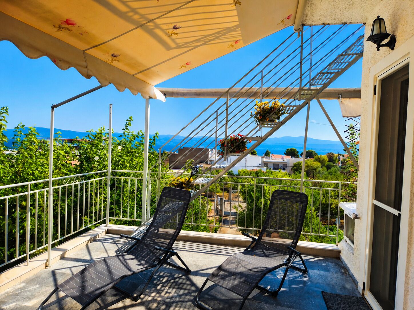 A balcony with two chairs, a metal railing, a covered area with a canopy, and a view of the sea and mountains.