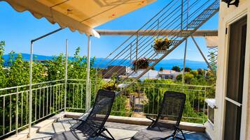 A balcony with two chairs, a metal railing, a covered area with a canopy, and a view of the sea and mountains.