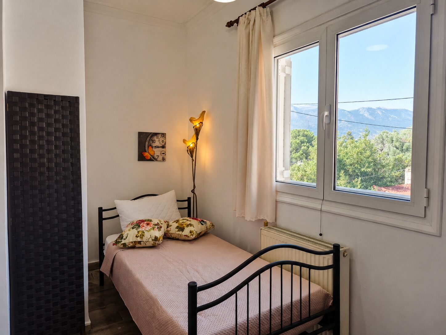 Une chambre à coucher comprenant un lit, une table de chevet, une horloge murale, une lampe sur pied et une fenêtre donnant sur des arbres et des montagnes.