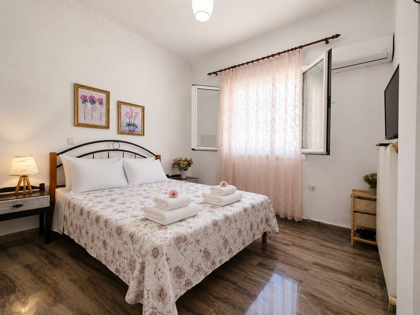 A bedroom with a bed, bedside table, lamp, two framed pictures, a window with curtains, and a wooden shelf.