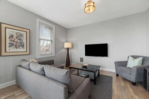 A modern living room with a grey sofa, a coffee table, and a television on the wall. - Skyline Luxury - Stylish 1BR with Panoramic Views (Pittsburgh)