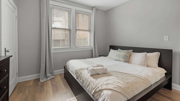 A bedroom with a bed, a dresser, and a window with blinds. - Skyline Luxury - Stylish 1BR with Panoramic Views (Pittsburgh)