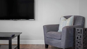 A living room with a flat-screen TV mounted on the wall, a grey armchair with a light blue pillow, and a small dark wooden table. - Skyline Luxury - Stylish 1BR with Panoramic Views (Pittsburgh)
