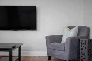 Ein Wohnzimmer mit einem an der Wand befestigten Flachbildfernseher, einem grauen Sessel mit einem hellblauen Kissen und einem kleinen dunklen Holztisch.