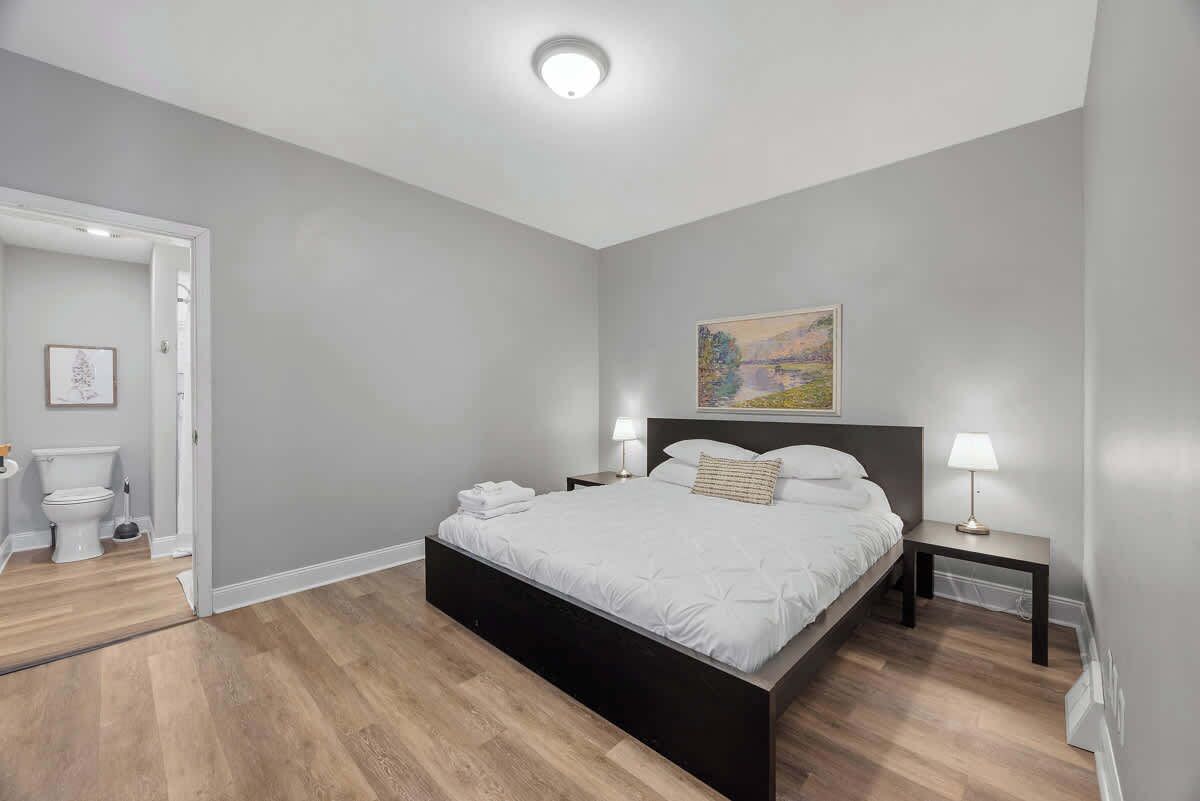 Une chambre à coucher moderne avec un grand lit, une table de chevet, une commode, un tableau au mur et une porte donnant sur une salle de bain.