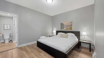 Une chambre à coucher moderne avec un grand lit, une table de chevet, une commode, un tableau au mur et une porte donnant sur une salle de bain.