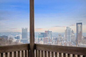 Vista urbana a través de una ventana con baranda metálica, mostrando varios edificios, incluyendo un rascacielos alto con una estructura particular en la parte superior.
