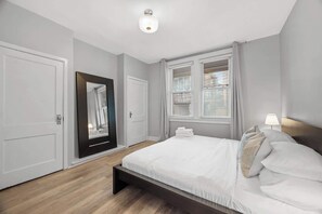 Een slaapkamer met een bed, nachtkastje, spiegel en een raam met jaloezieën.