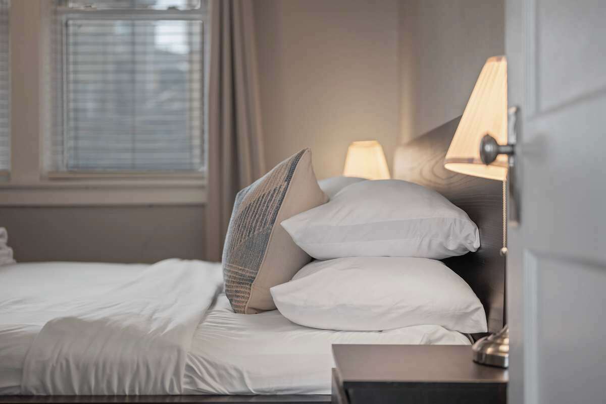 Un lit bien fait, avec du linge de lit blanc et un seul coussin à motifs, une lampe de chevet et une fenêtre avec des stores.