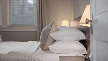 Un lit bien fait, avec du linge de lit blanc et un seul coussin à motifs, une lampe de chevet et une fenêtre avec des stores.