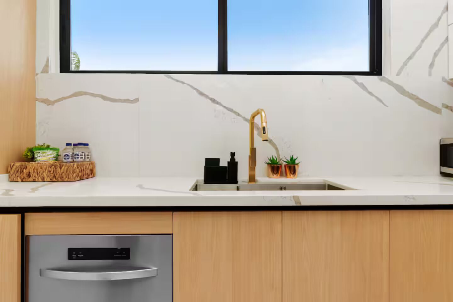 A modern kitchen with a white marble backsplash, a stainless steel dishwasher, wooden cabinets, a black faucet, and two potted plants on the countertop.
