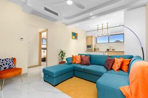 Un salon moderne avec un canapé d’angle, un petit fauteuil orange, un coin cuisine et une grande fenêtre donnant sur un beau paysage.