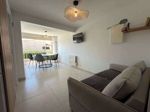 Un salon moderne comprenant un canapé, un coin repas avec des chaises et une table, un téléviseur fixé au mur et un luminaire suspendu au plafond.