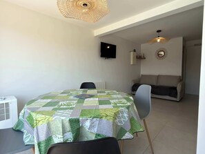 Ein Essbereich mit einem Tisch, bedeckt mit einer gemusterten Tischdecke, einem an der Wand montierten Fernseher und einer Couch im Hintergrund.