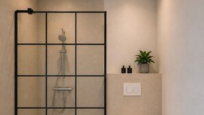 A modern bathroom with a glass shower enclosure, a toilet, a small shelf with toiletries, and a potted plant.