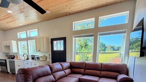 A modern living room with a sectional sofa, a ceiling fan, and large windows offering a view of the outdoors. - Modern New Cabin in East TN. Near Cherokee Lake and 1 hr to Pigeon Forge (Bean Station)