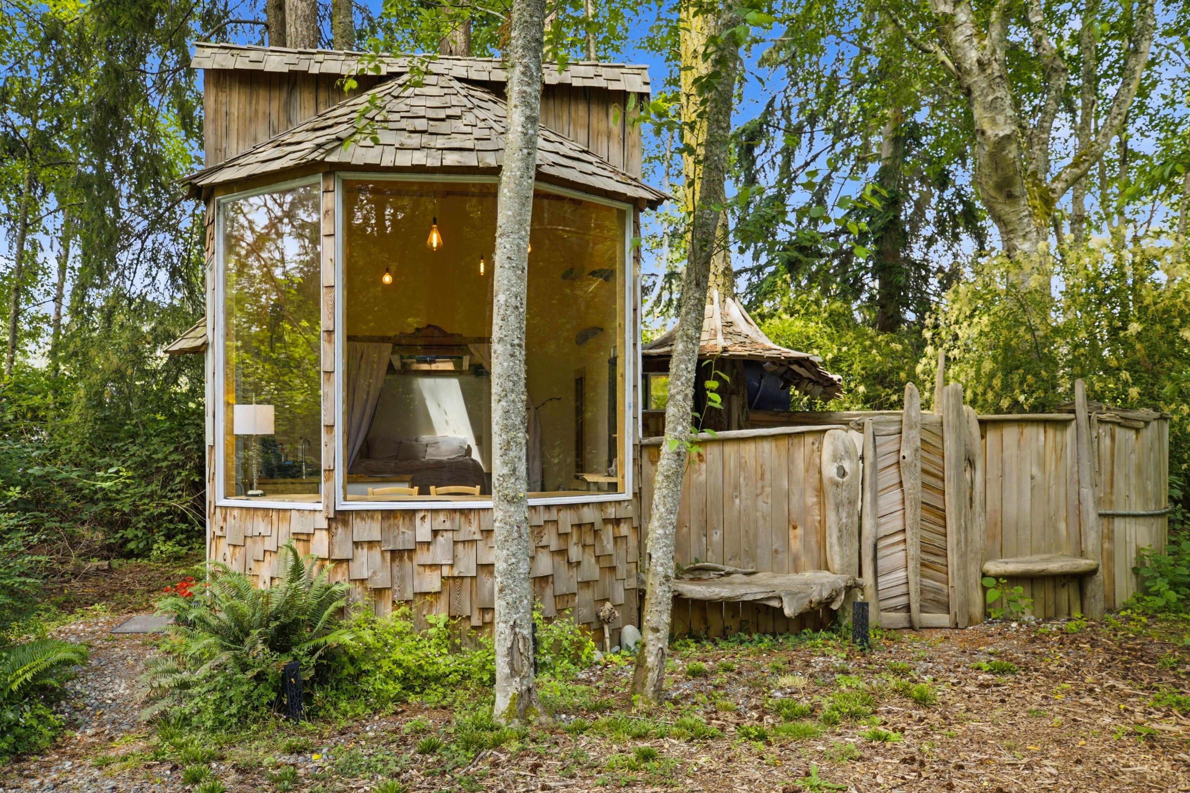 A wooden cabin with a large window, surrounded by trees and a wooden fence.