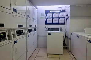 A laundry facility with multiple washing machines and dryers, a tiled floor, and a bulletin board with handwritten notes.