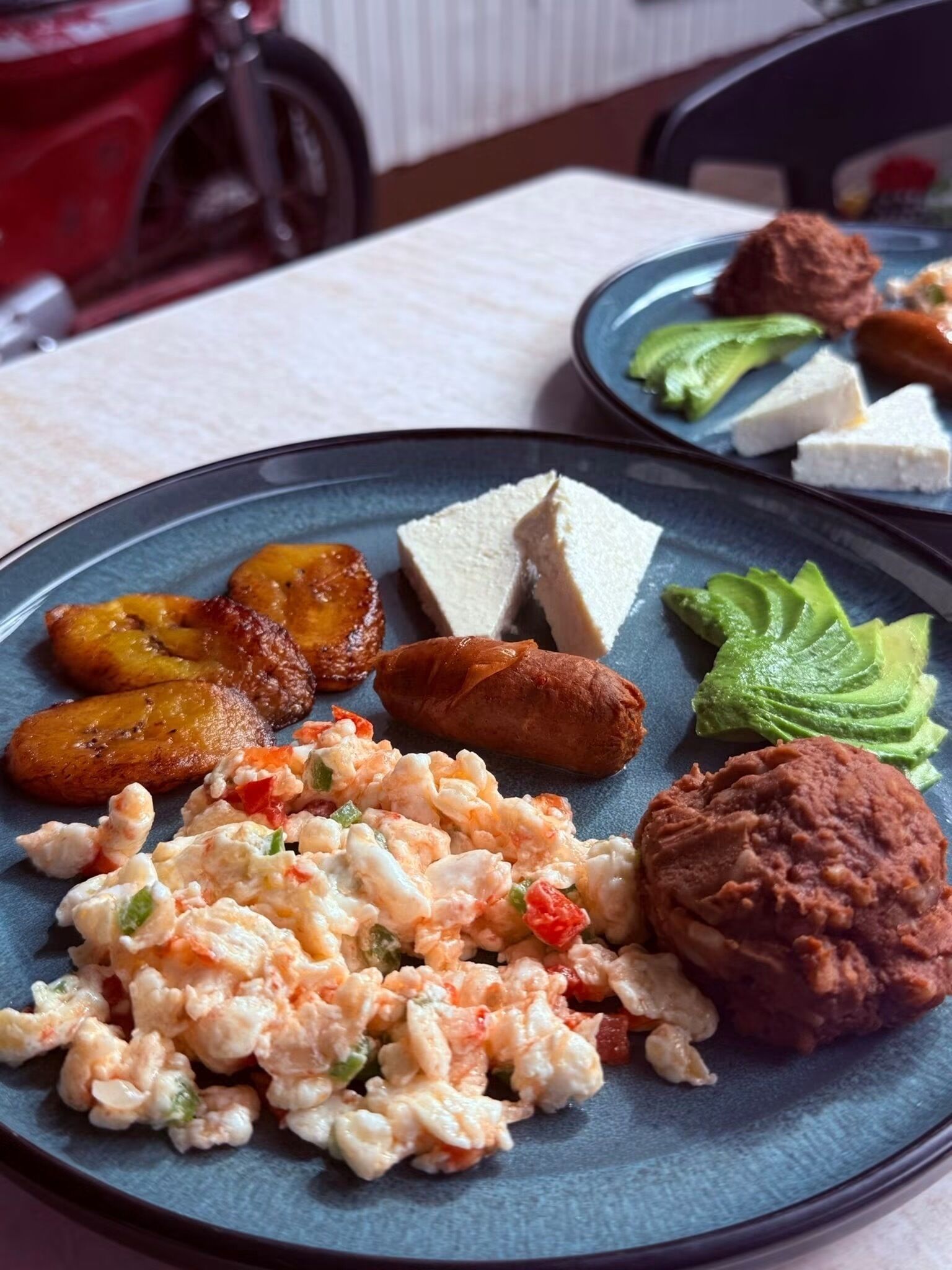 A plate with scrambled eggs, plantains, a sausage, avocado slices, and a meat patty.