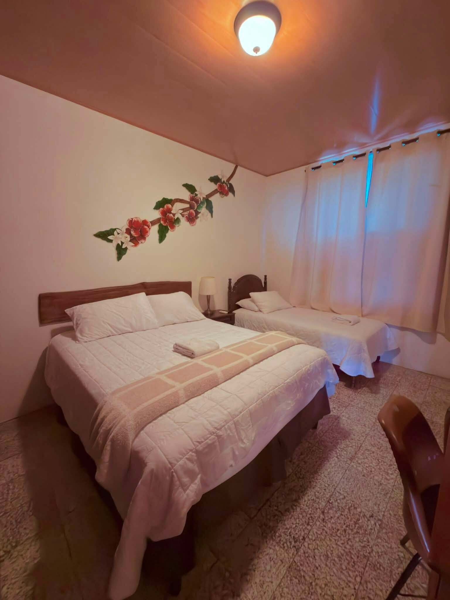 Ein Hotelzimmer mit zwei Betten, einem Nachttisch, einer Lampe, einem Sessel und einer Wand mit floralem Dekor.
