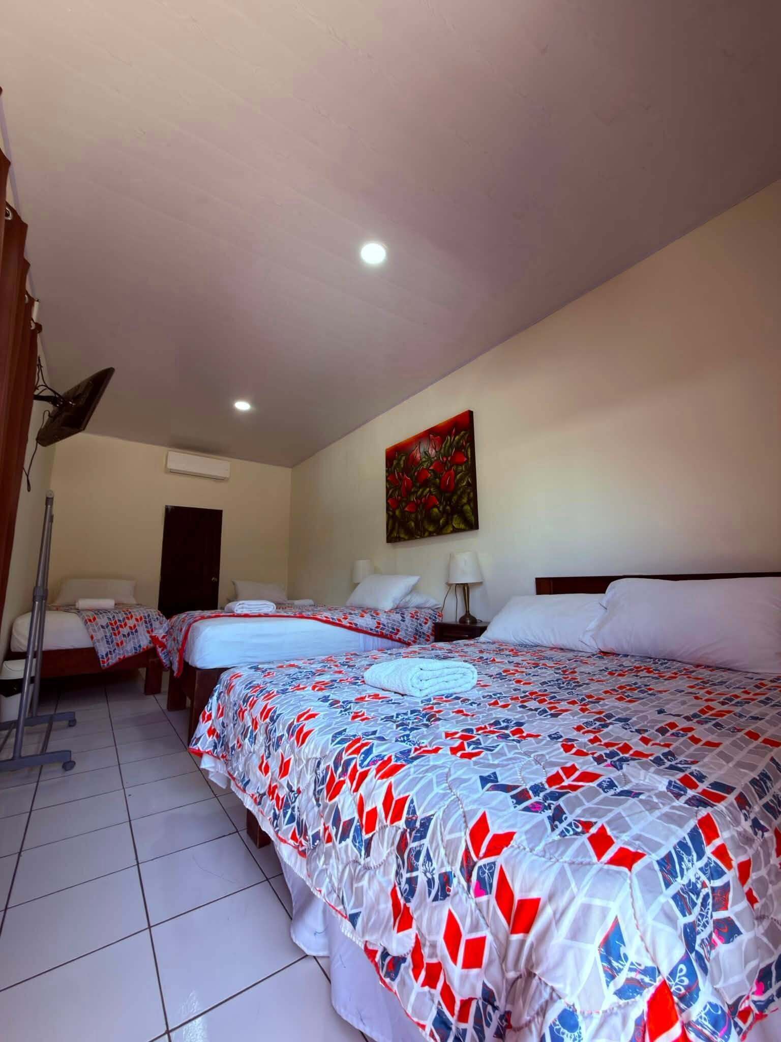 A hotel room with three beds, a painting on the wall, and a ceiling fan.