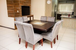 A dining area with a rectangular table and six chairs, a flat-screen TV mounted on a brick wall, and a kitchen area in the background.