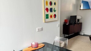 Ein modernes Wohnzimmer mit einem Glastisch, klaren Stühlen, einer dunklen Holzkommode und einem farbenfrohen, abstrakten Gemälde an der Wand.