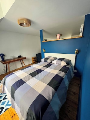 A bedroom with a checkered bedspread, a desk with a lamp, and a blue accent wall. - Blue Pink | Cozy Apartment in Championnet (Grenoble)