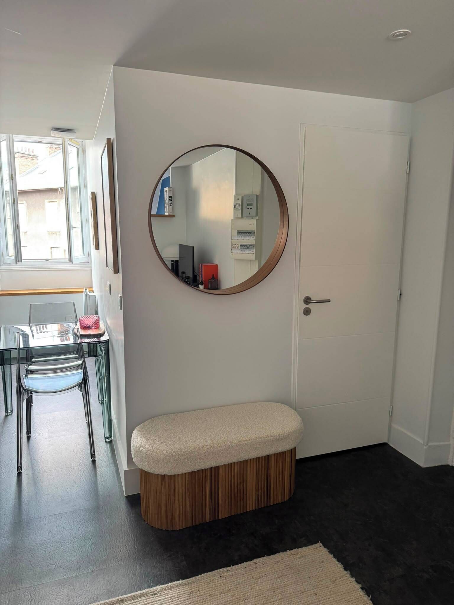 A room with a round mirror on the wall, a bench, a door, and a table with chairs.