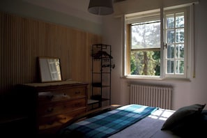A bedroom with a bed, a dresser, a mirror, a radiator, and a window with a view of trees. - Charming Villa with a Hot Tub in a Pine Forest (Talacchio)