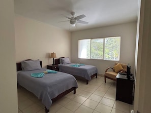 Ein Hotelzimmer mit zwei Betten, einem Schreibtisch, einem Stuhl, einem Deckenventilator und einem Fenster mit Jalousien.